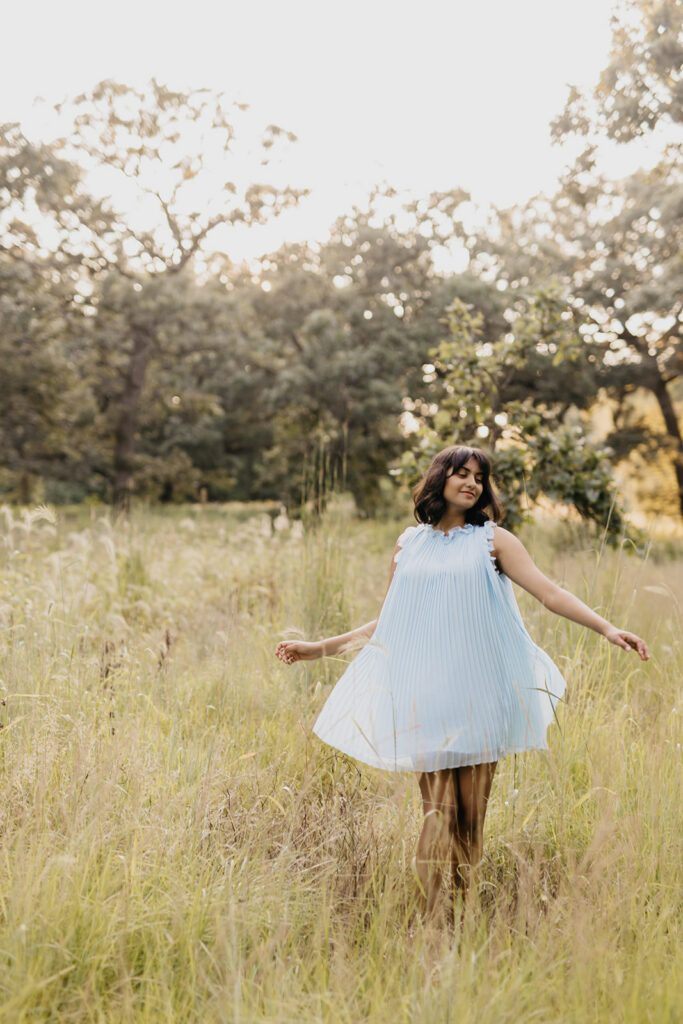 Senior girl in light blue pleated dress twirling in golden meadow during outdoor portrait session Minnesota