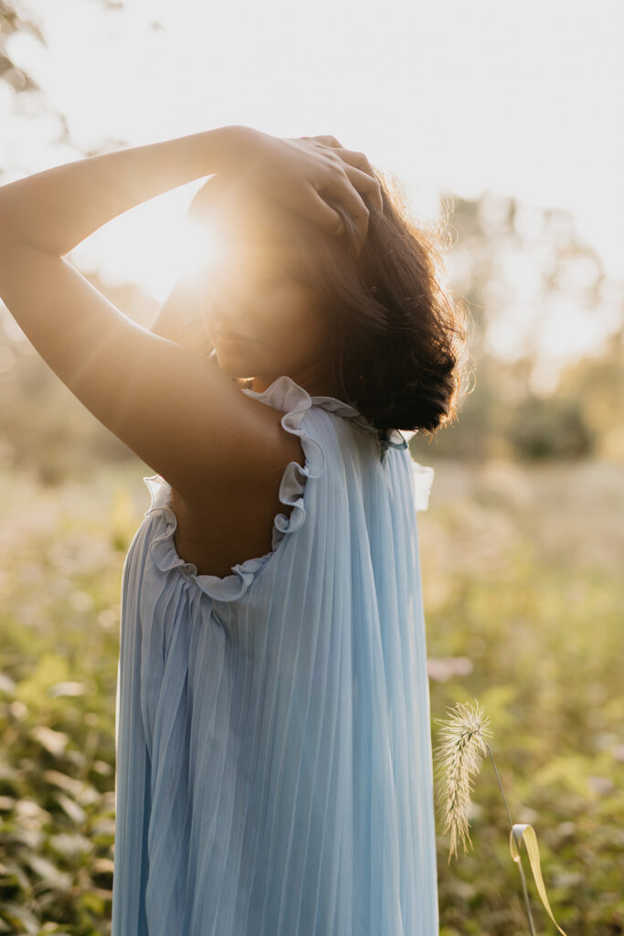 Senior girl in light blue dress with hand raised behind head backlit by golden sunset during senior photo session Minnesota