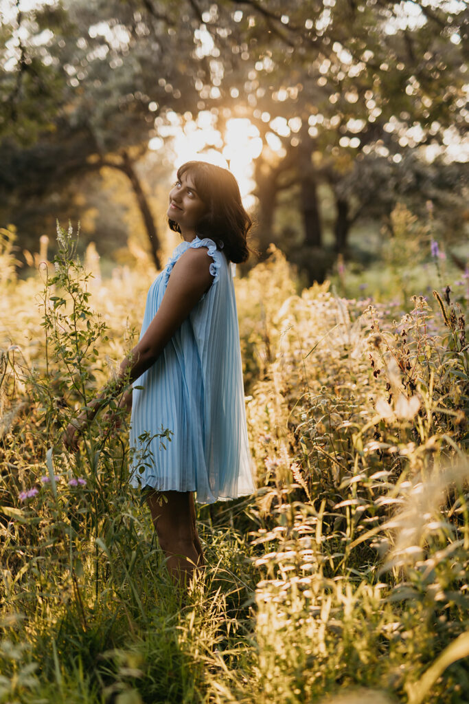 Senior girl in light blue dress standing in tall wildflower grasses looking up during golden hour senior portrait Minnesota