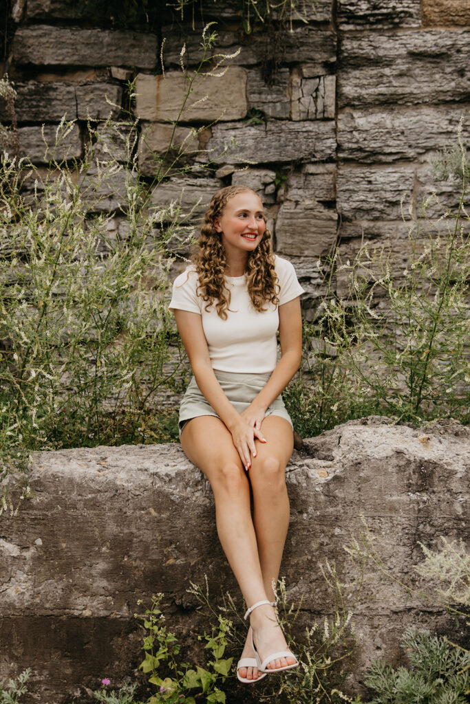 Senior girl in white top and sage skirt sitting on historic stone wall at Mill City Ruins senior photos Minneapolis