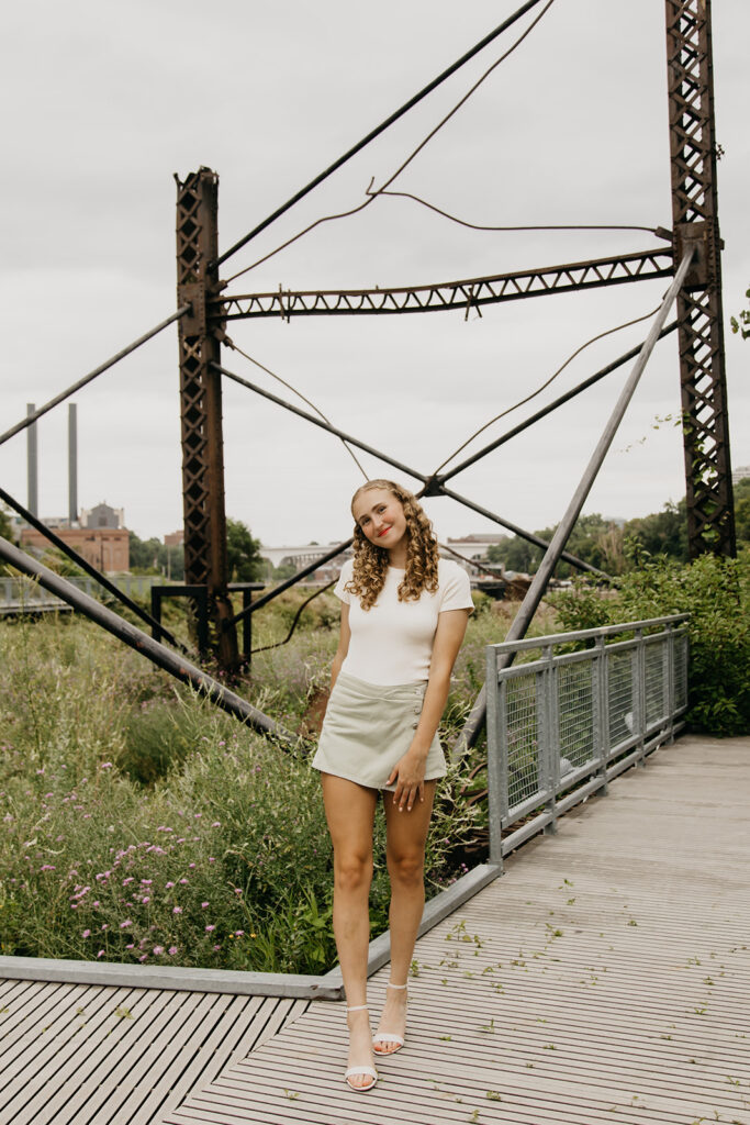 Senior girl in white top and sage skirt standing near industrial bridge at Mill City Ruins senior photo session Minneapolis