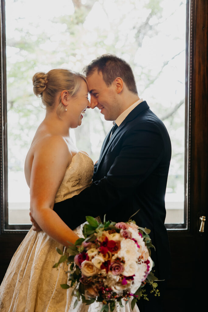 Bride and groom forehead to forehead in front of a large window with garden view, intimate wedding portrait Minnesota