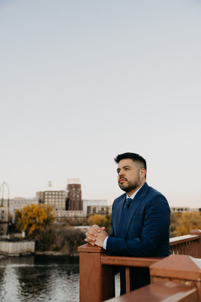 Brand photography Minneapolis — man in navy suit leaning on bridge railing overlooking the Mississippi River with Minneapolis skyline, by Mycah Bain Photography
