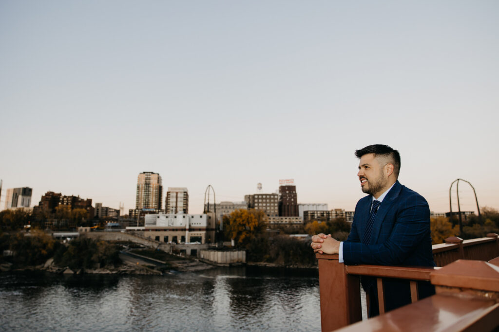 Professional brand photography Minneapolis — man in navy suit leaning on bridge railing with full Minneapolis skyline and Mississippi River in background, by Mycah Bain Photography
