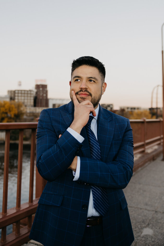 Professional brand portrait Minneapolis — man in navy suit with hand on chin in thoughtful pose on a bridge with city skyline, by Mycah Bain Photography