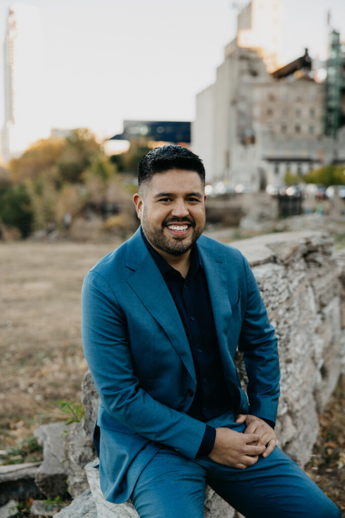 Professional brand portrait Minneapolis — man in navy suit with hand on chin in thoughtful pose on a bridge with city skyline, by Mycah Bain Photography
