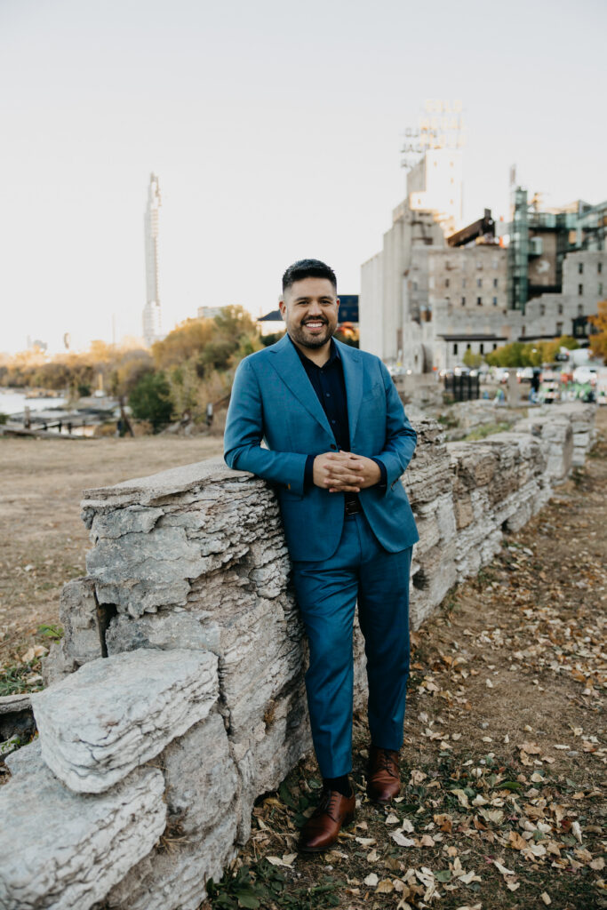 Brand portrait Minneapolis — man in teal suit leaning against historic stone wall smiling with Mississippi River and city skyline behind him, by Mycah Bain Photography