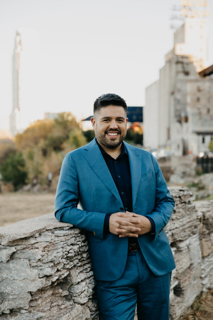 Brand photography Minneapolis — man in navy suit leaning on bridge railing overlooking the Mississippi River with Minneapolis skyline, by Mycah Bain Photography
