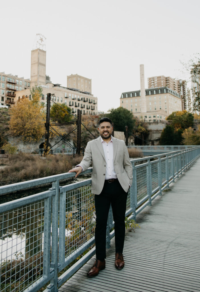Brand portrait Minneapolis — man in tan blazer standing full-length on riverfront walkway with Minneapolis skyline and fall foliage, by Mycah Bain Photography
