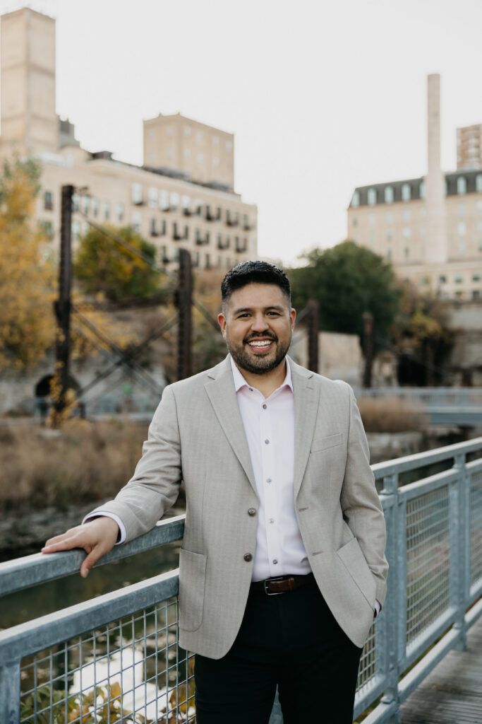 Brand photography Minneapolis — man in tan blazer standing on pedestrian bridge with Mill District skyline behind him, by Mycah Bain Photography