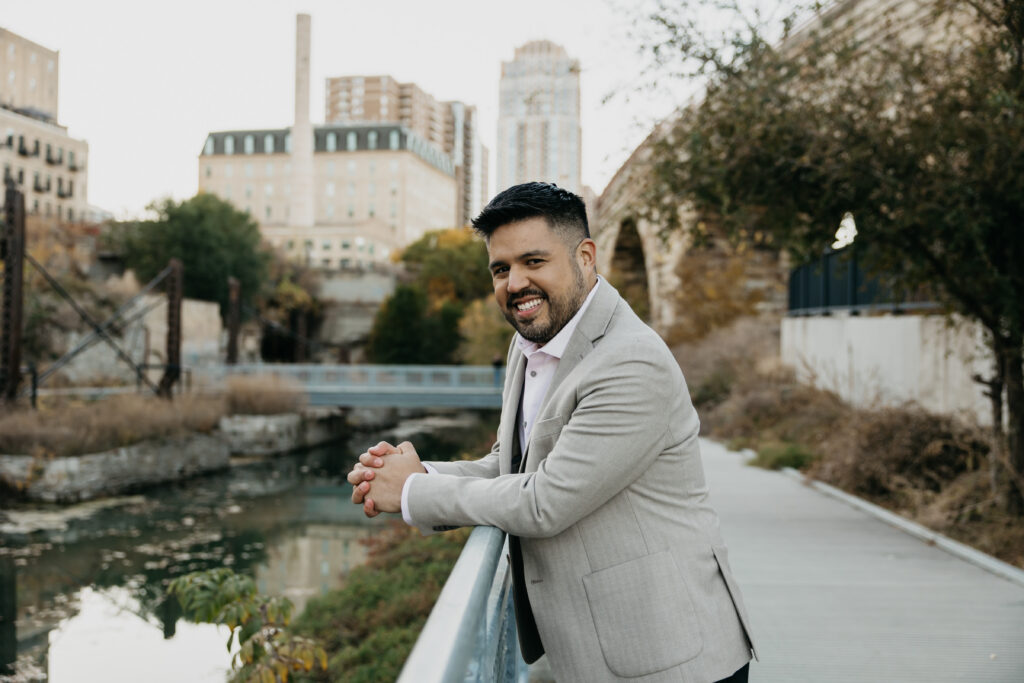 Professional brand headshot Minneapolis — man in tan blazer leaning on bridge railing smiling with Stone Arch Bridge and river in background, by Mycah Bain Photography