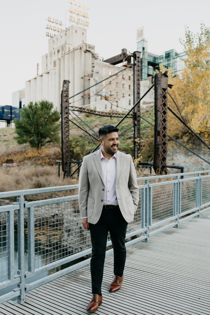 Brand photography Minneapolis — man in tan blazer walking on riverfront path with historic Stone Arch Bridge and Gold Medal Flour building in background, by Mycah Bain Photography
