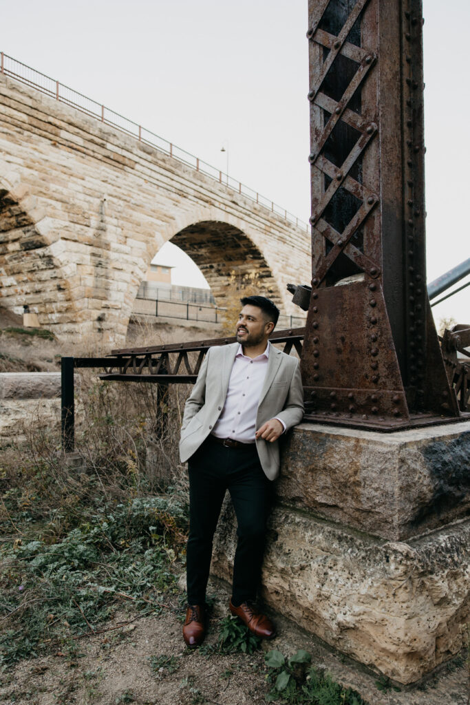 Professional brand portrait Minneapolis — man in tan blazer leaning against historic stone bridge pillar near the Mississippi River, by Mycah Bain Photography