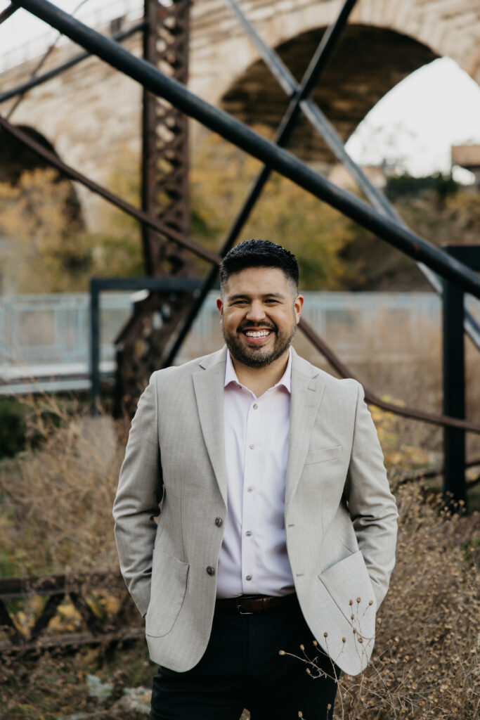 Brand headshot Minneapolis — man in tan blazer smiling confidently in front of historic Stone Arch Bridge arch, by Mycah Bain Photography
