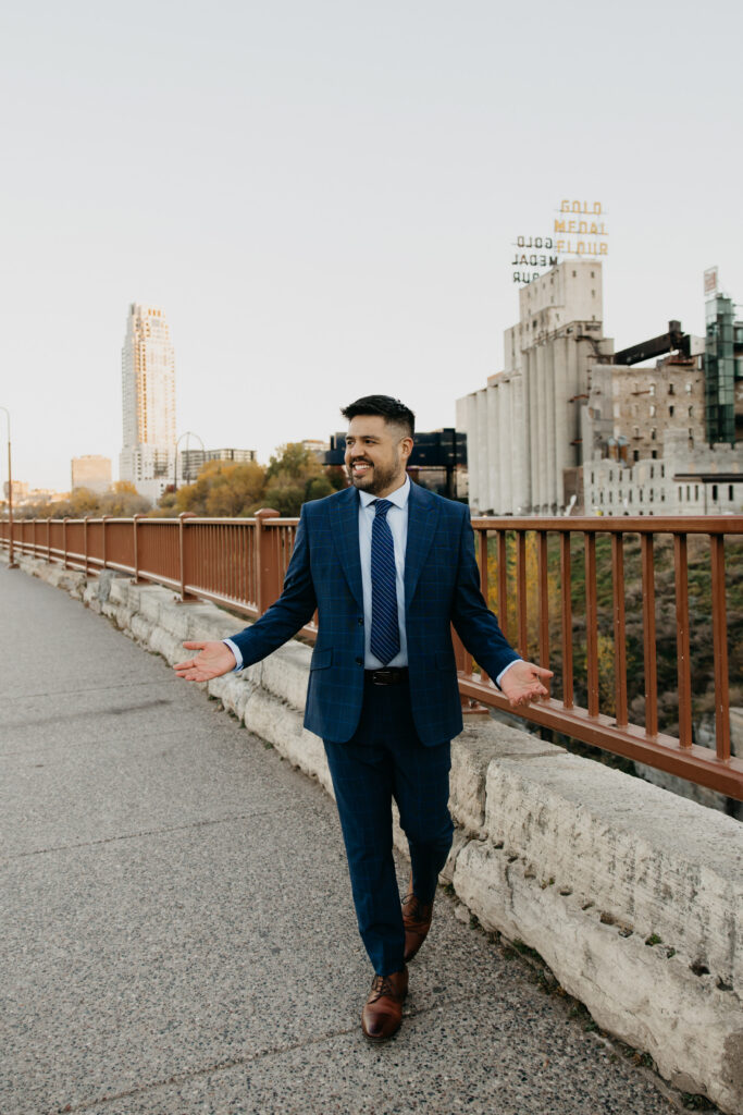 Brand photography Minneapolis — man in navy suit walking along bridge with Gold Medal Flour sign and skyline in background, by Mycah Bain Photography