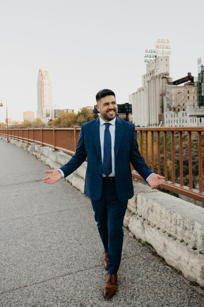 Brand photography Minneapolis — man in navy suit walking confidently along Stone Arch Bridge with arms open and Mill District in background, by Mycah Bain Photography