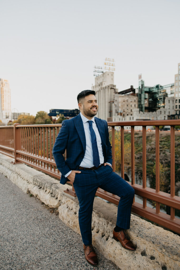 Professional brand portrait Minneapolis — man in navy suit sitting casually on bridge wall with Mill District skyline, by Mycah Bain Photography
