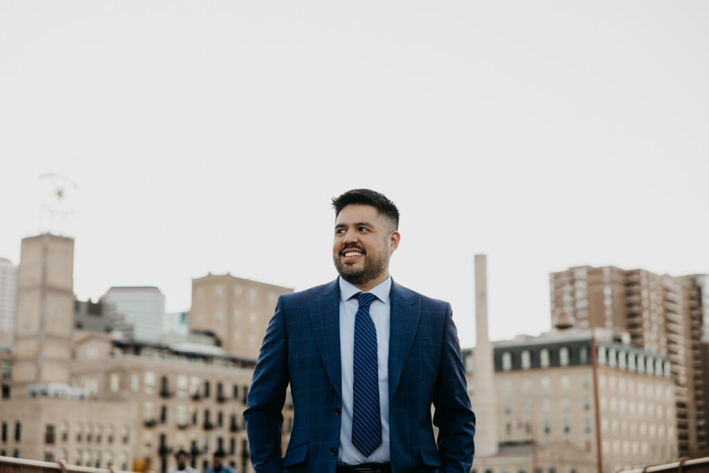 Brand headshot Minneapolis — man in navy suit smiling with hand in pocket against urban Minneapolis architecture, by Mycah Bain Photography