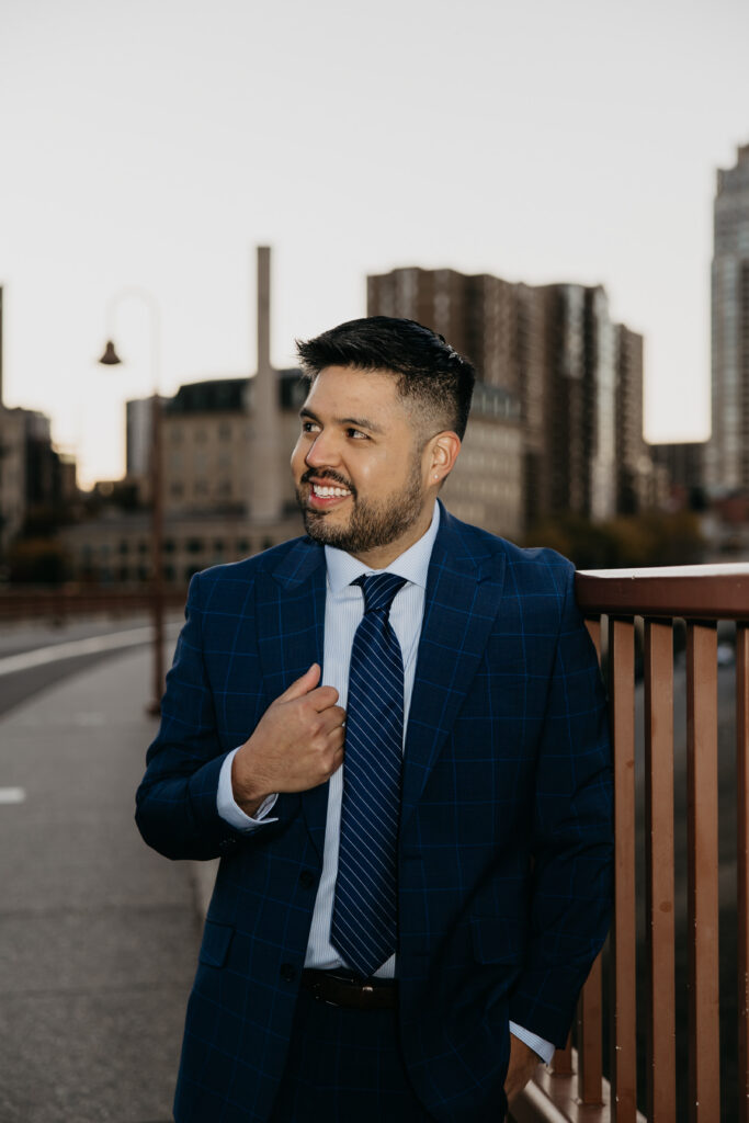 Professional brand headshot Minneapolis — man in navy suit smiling off-camera on a bridge with city skyline in background, by Mycah Bain Photography