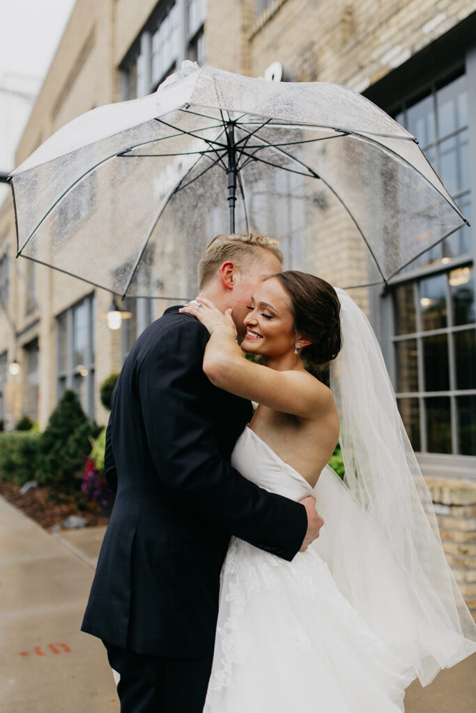 Bride and groom smiling and embracing in the rain under a clear umbrella, Minneapolis Warehouse District wedding