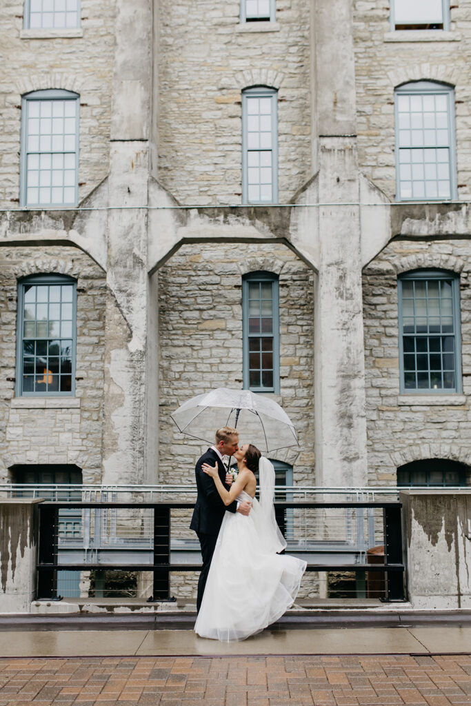 Bride and groom embracing under an umbrella in front of a historic stone building in the Minneapolis Warehouse District