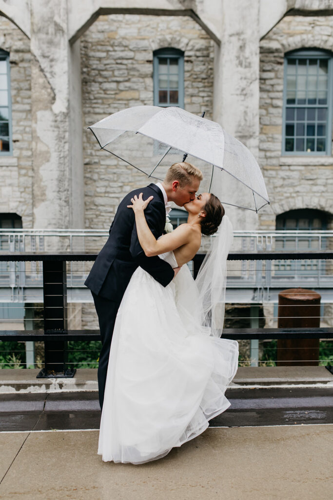 Bride and groom dipping kiss under a clear umbrella in the rain outside a historic stone building, Minneapolis wedding
