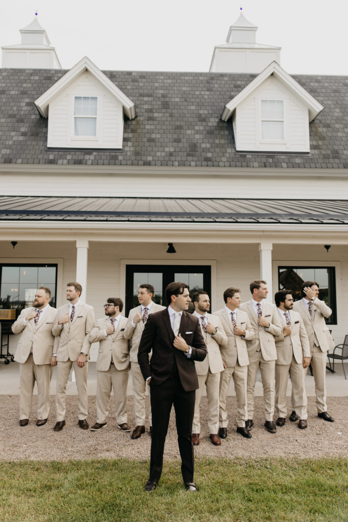 Groomsmen in tan suits lined up with groom in dark suit in front of a white farmhouse wedding venue Minnesota