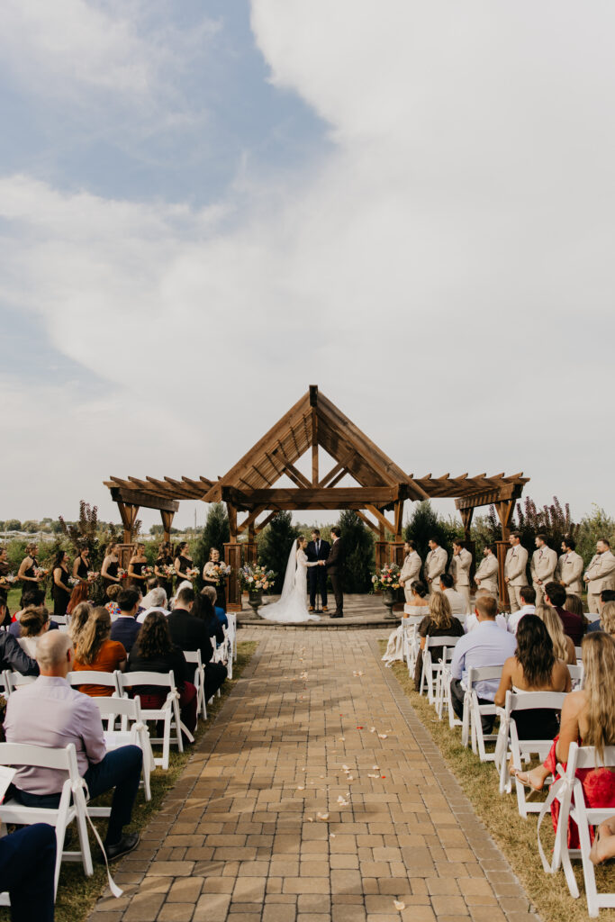 Wide ceremony aisle view at an outdoor Minnesota wedding with wooden pergola and guests seated on white chairs