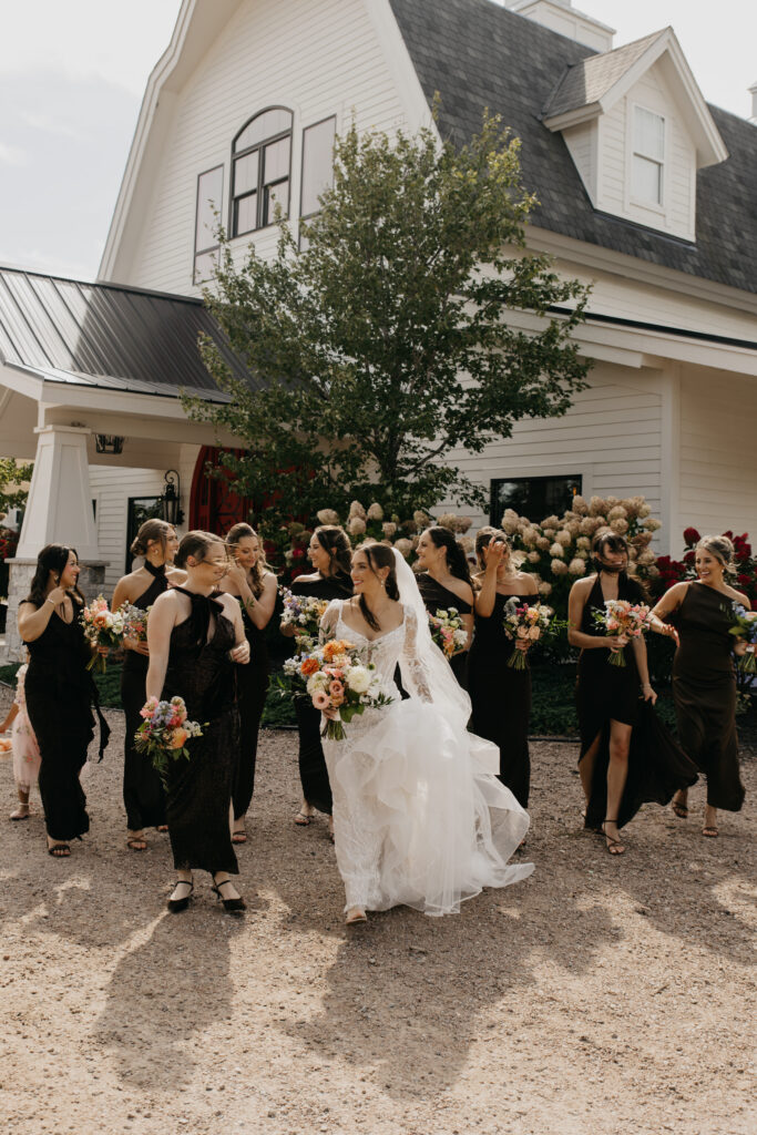 Bridesmaids in black dresses walking with bride in front of a white farmhouse wedding venue in Minnesota