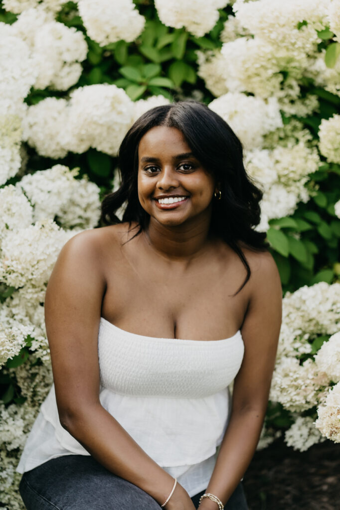 Senior girl in white strapless top seated in front of full white hydrangea blooms garden senior photo session Minnesota