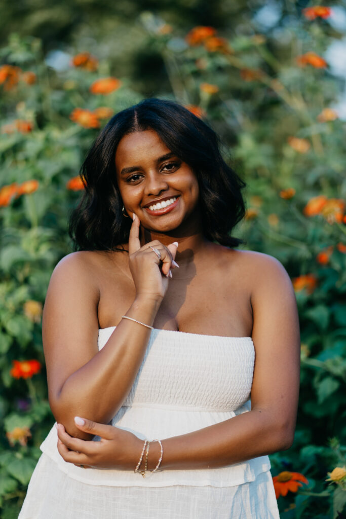 Senior girl in white smocked tube top smiling in front of vibrant orange flowers during senior portrait session Twin Cities