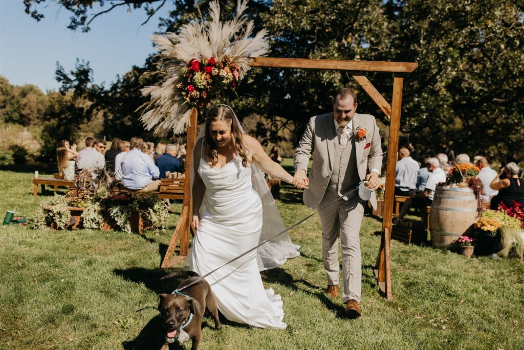 Bride and groom recessional with their dog under a wooden arch at an outdoor fall wedding in Minnesota, captured by Mycah Bain Photography
 