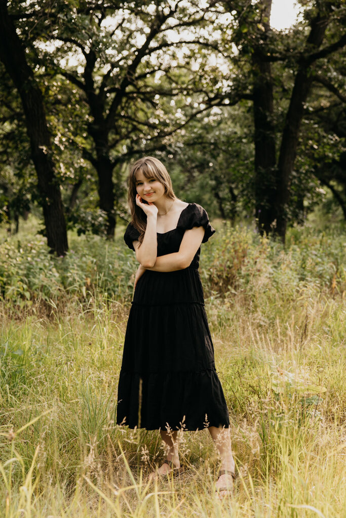 Senior girl in black dress standing in shaded woodland area during outdoor senior photo session Twin Cities Minnesota
