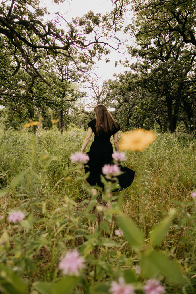 Senior girl in black dress walking away through wildflower field with pink and yellow blooms senior portrait Minnesota