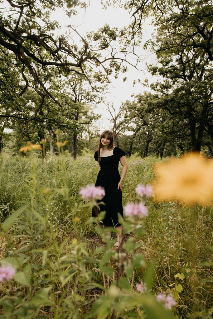 Senior girl in black dress standing among wildflowers shot through foreground flowers during senior photo session Twin Cities