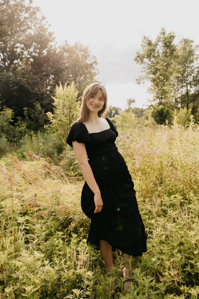 Senior girl in black puff sleeve dress standing in tall grass meadow smiling during outdoor senior portrait session Minnesota