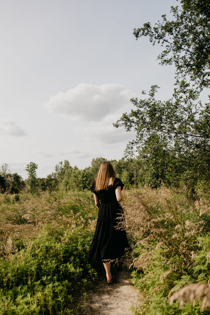 Senior girl in black dress walking away down nature path through summer greenery during senior portrait session Minnesota