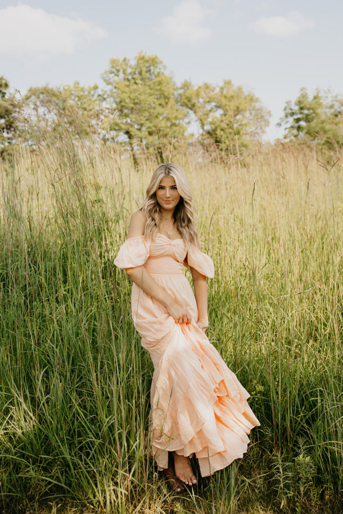 Senior girl in peach off-shoulder maxi dress standing in tall prairie grass during outdoor senior portrait session Minnesota