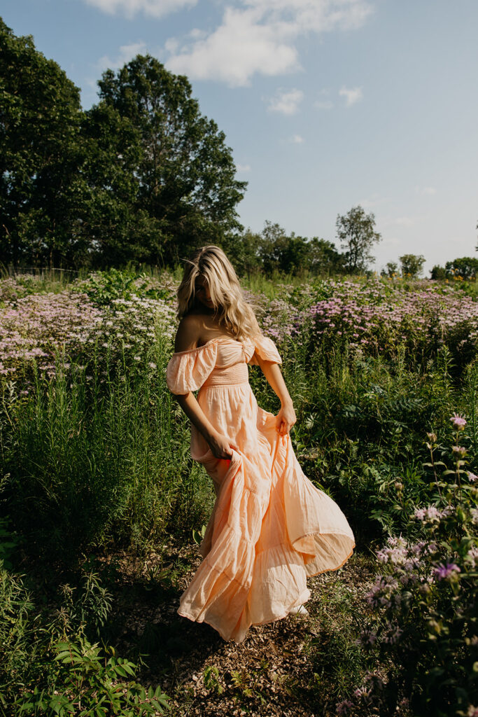 Senior girl in peach maxi dress walking away through wildflower meadow during summer senior photo session Twin Cities