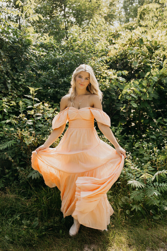Senior girl in peach off-shoulder maxi dress standing in lush green garden setting during senior portrait session Minnesota