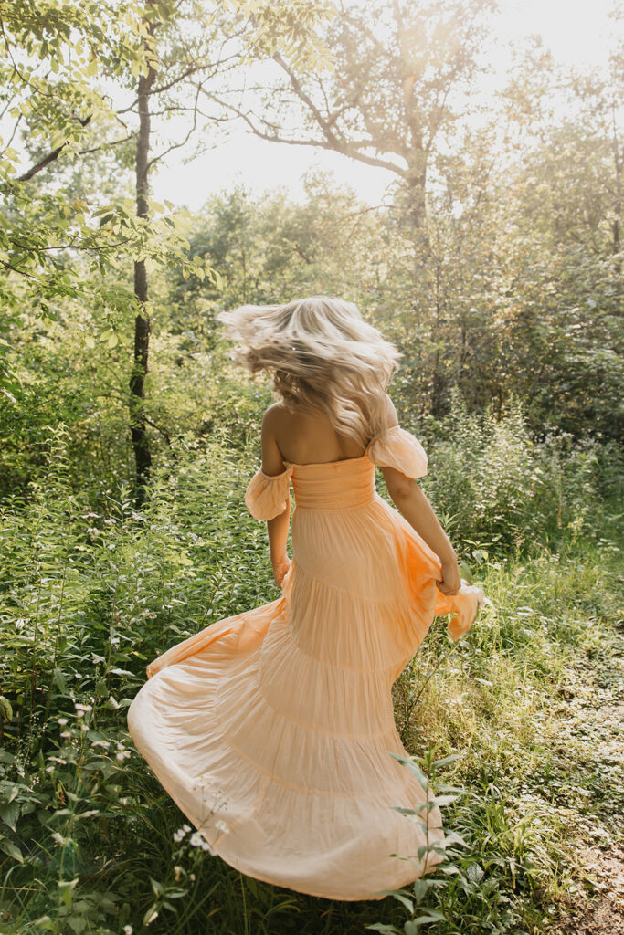 Senior girl in peach maxi dress mid spin from behind with hair flying in sunlit forest during senior photo session Minnesota