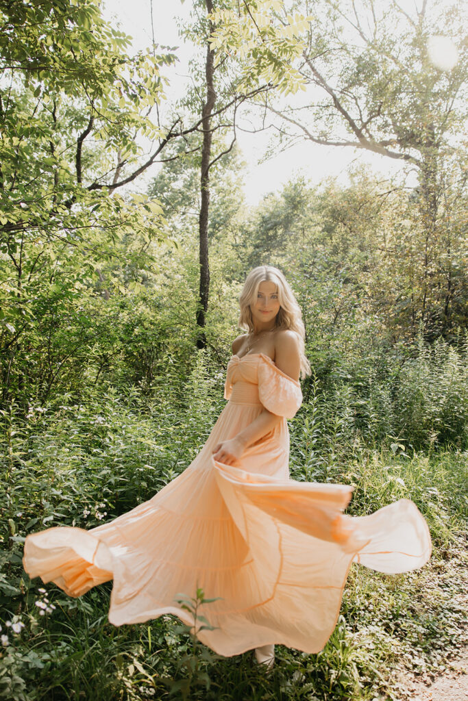 Senior girl in peach off-shoulder maxi dress spinning with skirt swirling in dappled forest light senior portrait Twin Cities