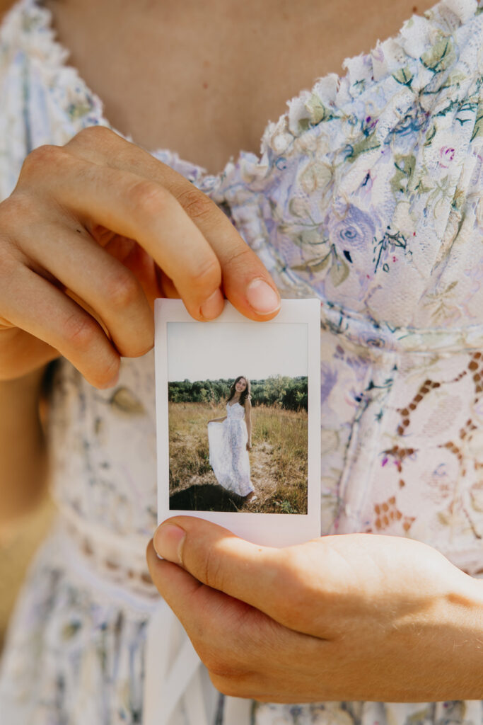 Senior girl in floral dress holding Polaroid photo of herself in white dress in a field during senior portrait session Minnesota