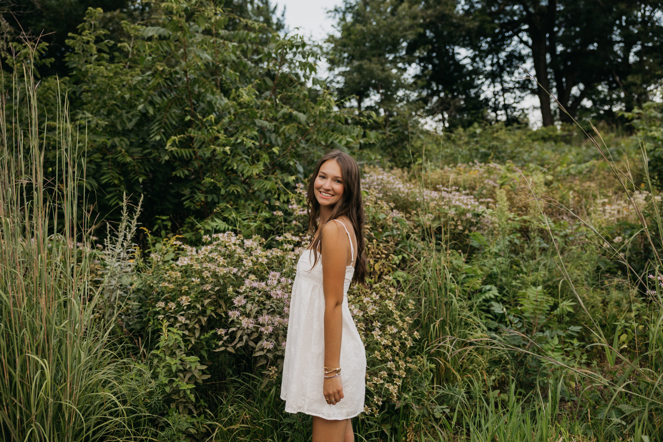 Senior girl in white eyelet dress smiling over shoulder in wildflower garden during outdoor senior portrait session Minneapolis Minnesot