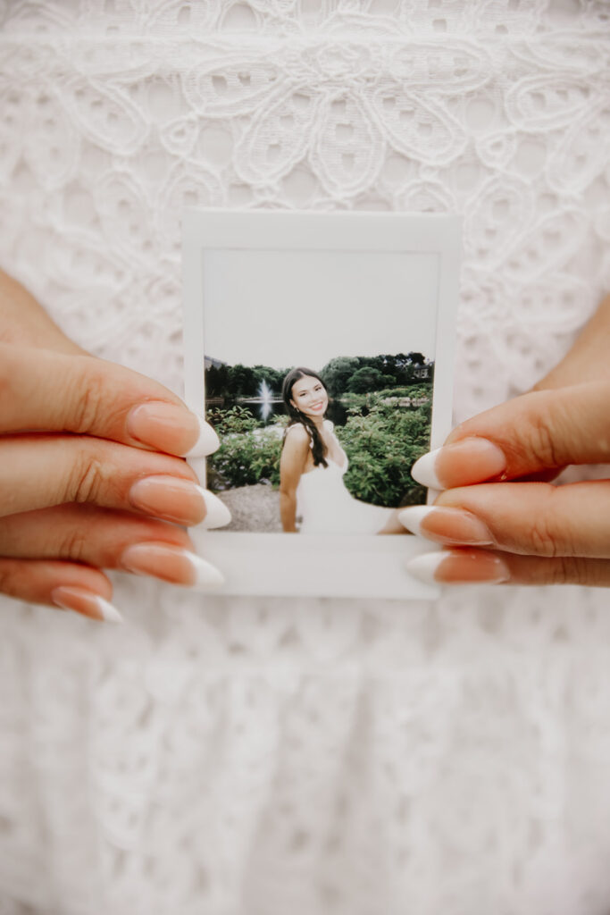 Senior girl in white eyelet lace top holding Polaroid photo of herself in garden setting with French tip nails Twin Cities senior photos