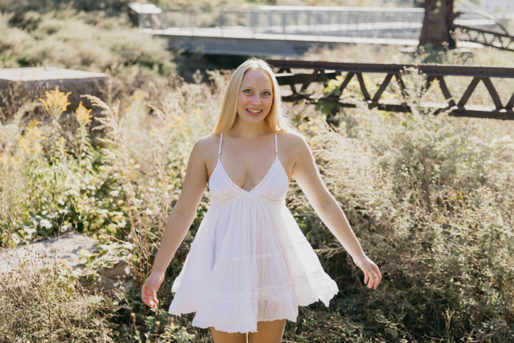 Senior girl in white spaghetti strap mini dress smiling while walking through tall golden grass near Mill City Ruins Minneapolis Minnesota