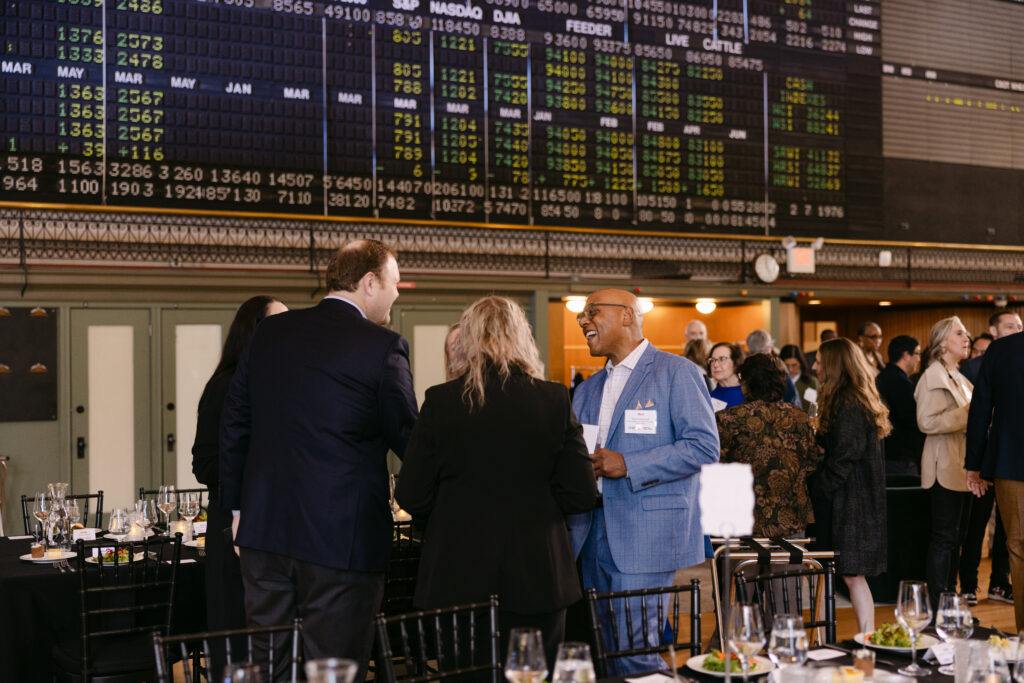 Two MPR gala guests laughing in conversation in front of the historic Grain Exchange stock ticker board — corporate event photography Minneapolis