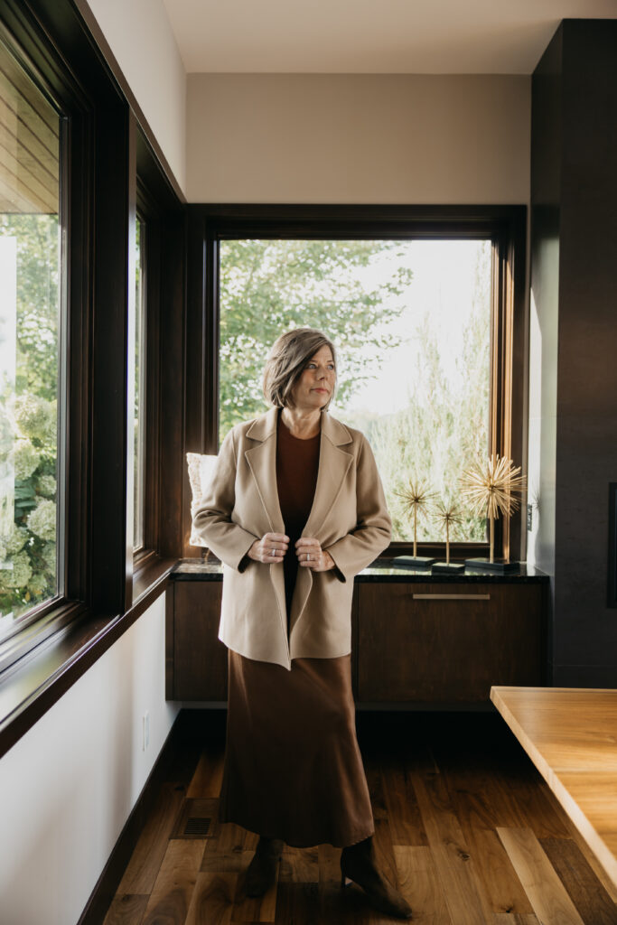 Dr. Melissa Polusny board-certified psychologist standing by dark-framed windows in camel coat and brown skirt during Minneapolis brand photography session by Mycah Bain Photography