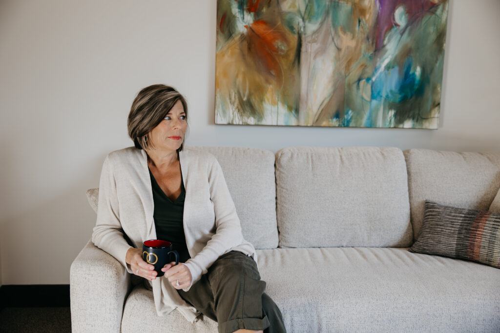 Dr. Melissa Polusny Minneapolis psychologist seated on cream sofa holding ARMOR mug with abstract art on wall — brand photography session by Mycah Bain Photography