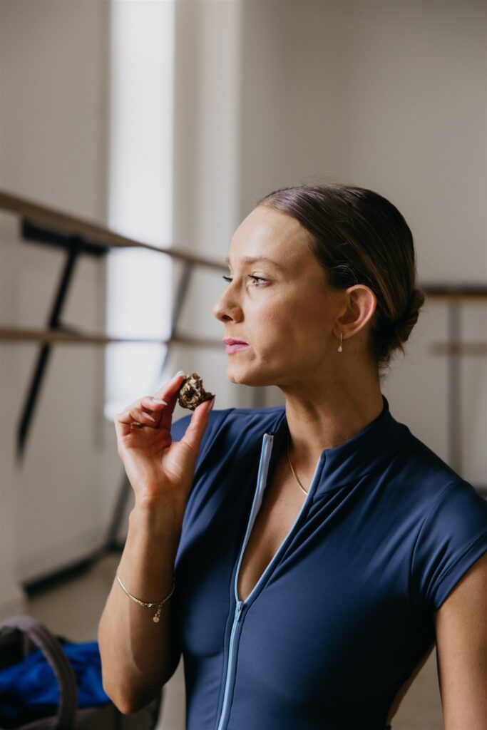 Lili Pfeifer of The Barbell Ballerina in navy unitard eating a bite of food with thoughtful expression during nutrition brand photography Minneapolis session by Mycah Bain Photography